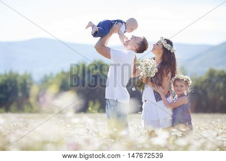 Happy family of four,mother,father,older sister and little brother spend time together in the highlands on a field of blooming white daisies in summer in the fresh air,mother and daughter wearing wreaths of flowers-white daisies