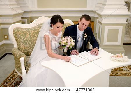 Beauty bride and handsome groom are registering the marriage. Wedding couple is marrying. Beautiful model girl in white dress and in lace veil. Man in suit. Female and male portrait. Cute lady and guy