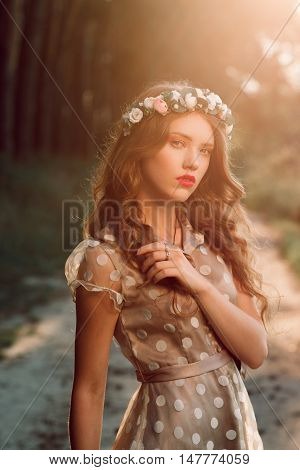 Attractive girl in wreath posing in forest. Beautiful fashion woman meet sunset in country, free space. Natural hair care, unity with nature concept