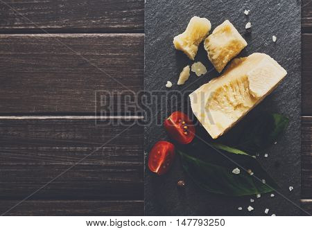 Cheese delikatessen closeup on black stone desk at wooden surface background. Parmesan pieces decorated with basil and cherry tomatoes, top view image with copy space, soft toning