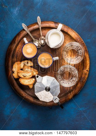 Coffee espresso in cups with italian cantucci, cookies and milk in jug on wooden serving round board over dark blue painted plywood background, top view, vertical composition