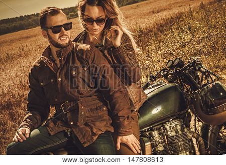 Young, stylish cafe racer couple on the vintage custom motorcycles in a field.
