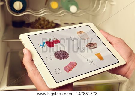 young man making his shopping list in his tablet computer, with pictures of different products shot by myself in its screen, in front of the fridge
