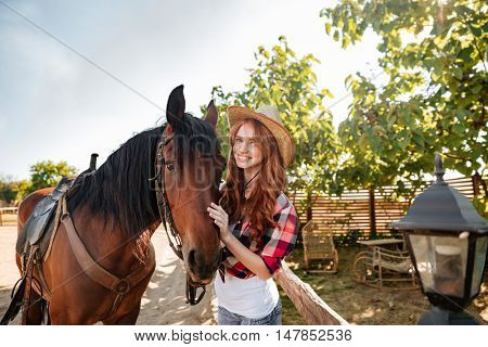 Cheerful beautiful young woman cowgirl standing with her horse in village