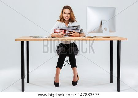 Full length portrait of a happy businesswoman holding folders while sitting at the office desk isoltaed on the white background