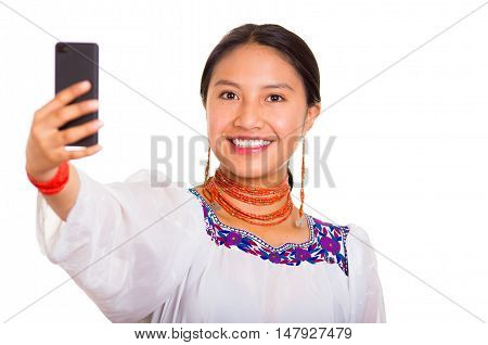 Beautiful young woman standing wearing traditional andean blouse and red necklace, holding mobile phone taking selfie while smiling happily, white studio background.