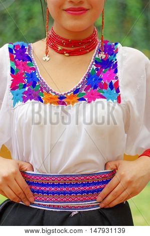 Closeup beautiful hispanic woman wearing black skirt, traditional andean white blouse with colorful decoration around neck and waist, matching red necklace, bracelet, ear ring, garden background.