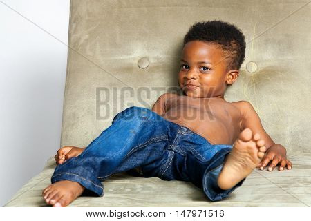 A precocious young African American boy reclines on a couch that he has colored on with a yellow crayon. His pants and face are a bit messy and he is smirking.