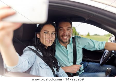 road trip, leisure, couple, technology and people concept - happy man and woman driving in car and taking selfie with smartphone