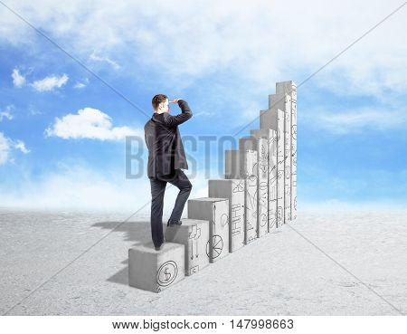 Back view of young businessman in suit looking into the distance while standing on abstract concrete chart bars with business doodles on sky background. Success and financial growth concept