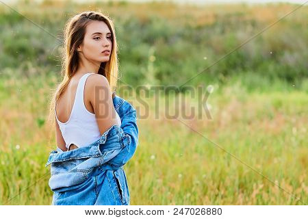 Happy Smiling Young Girl Wearing Blue Jeans Jacket And White Dress Is Standing In The Field