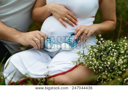 Cheerful Young Couple With Baby Shoes Sitting On A Flower Field With White Daisies. Pregnancy Concep