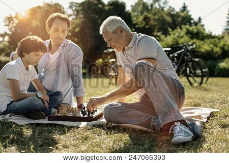 I Love Chess. Concentrated Grey-haired Grandpa Sitting On The Grass And Playing Chess With His Small