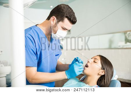 Male Dentist Examining Teenage Girl With Dental Mirror And Carver At Clinic