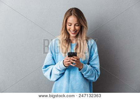 Pretty young blonde girl in blue sweatshirt using mobile phone isolated over gray background