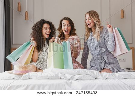 Photo of young beautiful women 20s wearing dresses unpackaging colorful shopping bags while resting on luxury bed in hotel room during bachelorette party