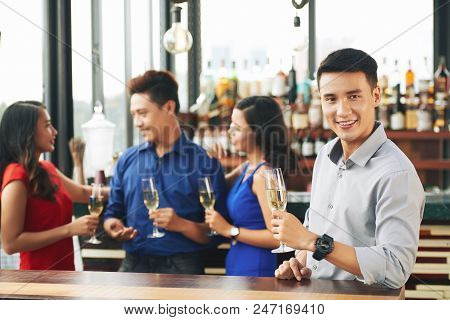 Portrait Of Cheerful Vietnamse Young Man With Glass Of Champagne, His Friends Are In Background