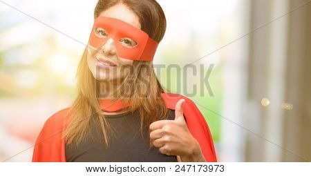Middle age super hero woman wearing red mask and cape smiling broadly showing thumbs up gesture to camera, expression of like and approval