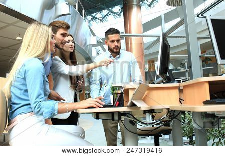 Group Of Young People In Casual Wear Sitting At The Office Desk And Discussing Something While Looki