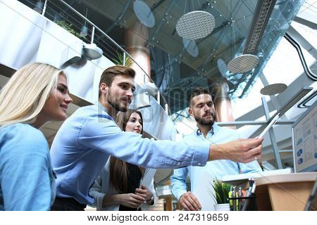 Group Of Young People In Casual Wear Sitting At The Office Desk And Discussing Something While Looki