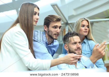 Group Of Young People In Casual Wear Sitting At The Office Desk And Discussing Something While Looki