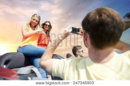 leisure, road trip, travel and people concept - happy friends taking picture by smartphone in convertible car over sky background