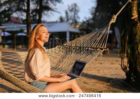 Female Freelancer Working With Laptop And Sitting On Sand In Wicker Hammock. Concept Of Modern Techn