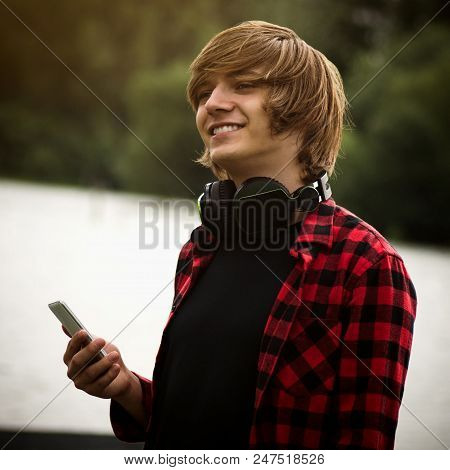 The Smiling Teenage Boy Or High Schooler Boy With Headphones Is Holding Mobile Phone In Hand Outdoor