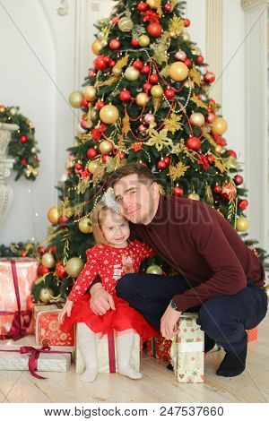 Little Pretty Daughter Wearing Red Dress Hugging Father Near Christmas Tree And Presents. Concept Of