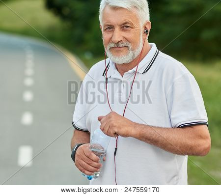 Portrait Of Senior Sportman Keeping Water Bottle During Morning Scamper. Looking At Camera, Listenin