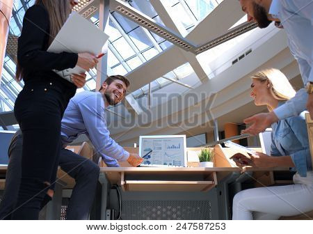 Group Of Young People In Casual Wear Sitting At The Office Desk And Discussing Something While Looki