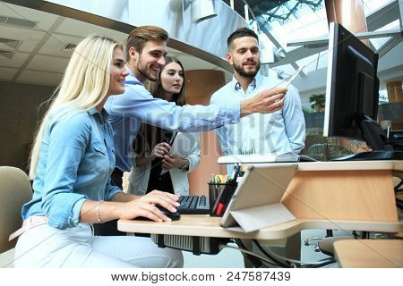 Group Of Young People In Casual Wear Sitting At The Office Desk And Discussing Something While Looki