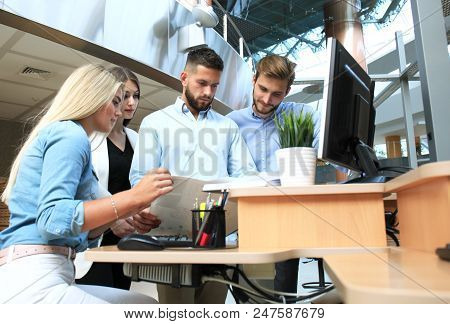 Group Of Young People In Casual Wear Sitting At The Office Desk And Discussing Something While Looki