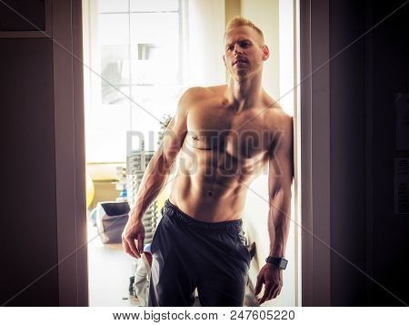 Handsome Muscular Shirtless Young Man Standing In A Gym, Looking At Camera, Leaning On Open Door