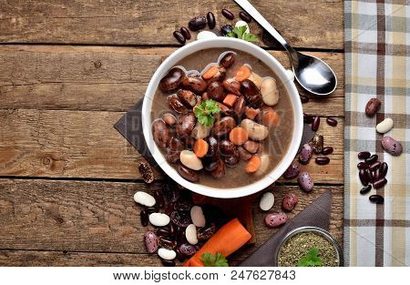 Top View Of Full Bowl Of Hot Bean Soup With Large Beans On Cutting Board, Carrots, Parsley, Marjoram