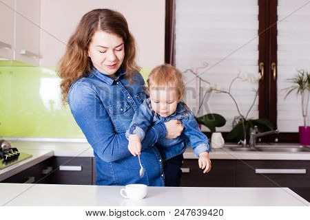 Cute Little Baby In The Arms Of His Mother In Their Kitchen. Small Boy Trying To Stirr Coffee With S