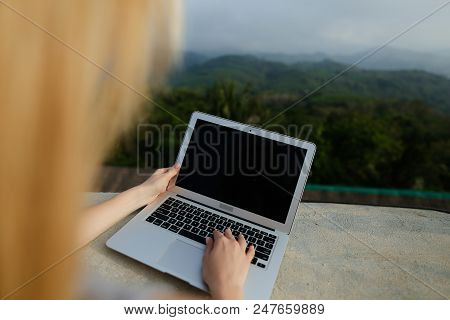 Young Blonde Girl Working With Laptop In Green Mountains Background, Wearing Grey Shirt. Concept Of 