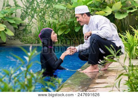 Muslim woman or girl in swimming pool in tropical garden wearing Burkini halal swimwear greeting her husband