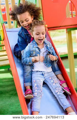 Two Excited Screaming Multiethnic Little Kids Sliding Down From Hill At Playground
