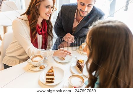 High-angle view of a happy couple sharing cakes while sitting at table with their mutual friend in a trendy coffee shop
