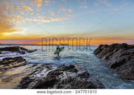 Surfer Standing On The End Of A Rock With Incoming Ocean Surf, And A Colourful Sunset At Currumbin R