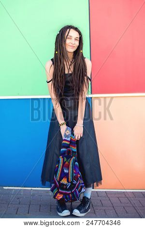 Beautiful Slim Girl With Dreadlocks, A Rastaman, In A Black Dress And Sneakers, Stands Against The B