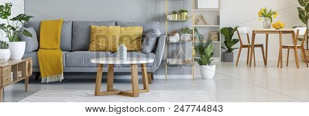 Real Photo Of A Grey Sofa With Yellow Pillows Standing Behind A Table And Next To A Shelf With Ornam