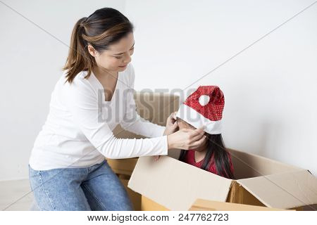 Woman Trying To Put Santa Claus Hat To Her Kid. Asian Girl Playing In The Box At Home. Kid Sitting I