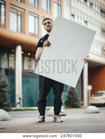 Young Attractive Man Holding Blank Canvas At The Street And Looking At The Viewer. Red Building On T