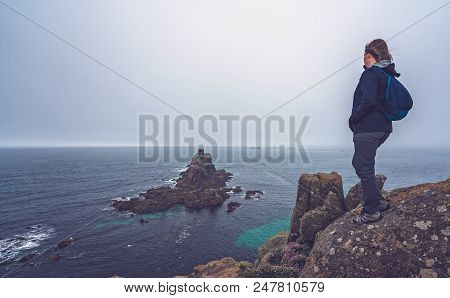 Lonely Tourist Dressed In Waterproof Jacket Standing On The Top Of A Cliff And Admiring The Rocky Fo