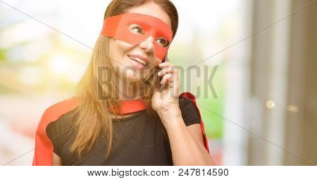 Middle age super hero woman wearing red mask and cape happy talking using a smartphone mobile phone