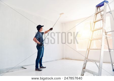 Asian Rear View Of A Male Painter Drawing A Wall With Paint Roller And A Separate Tank From A Large 