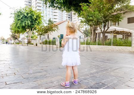 Back View Cute Little Blondy Toddler Girl In White Dress Standing Along On The Alley In The City Par