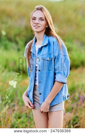Happy Smiling Young Girl Wearing Blue Jeans Jacket And Shorts Is Standing In The Field